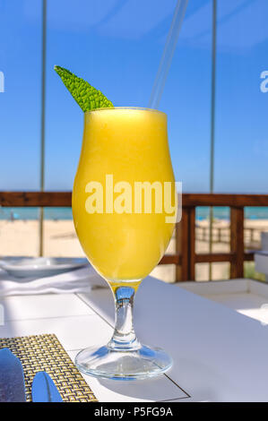 yellow mango smoothie fruit drink in a glass with a mint leaf and a straw on a table near a beach Stock Photo