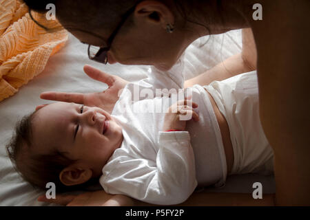 Baby laying on the bed smiling at his mom who is leaning over him. Stock Photo