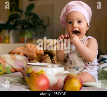 Baby girl dressed as a cook with a chef's cap on her head, holds a wooden spoon in front of a cooking pot and a few apples with some toys in the backg Stock Photo