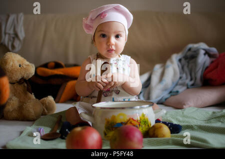 Baby girl dressed as a cook with a chef's cap on her head, holds a wooden spoon in front of a cooking pot and a few apples with some toys in the back. Stock Photo