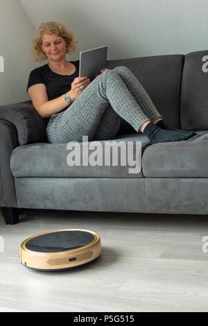 Middle aged woman is steering her vacuum cleaning robot using a digital tablet while resting on the sofa Stock Photo