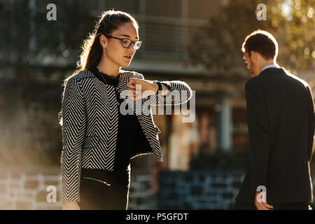 Woman looking at her wrist watch while commuting to office in the morning. Woman going to office checking time while walking on street with sun flare  Stock Photo