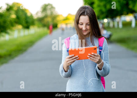 Girl schoolgirl. Summer in nature. Hands holding a tablet behind his backpack. Free space for text. The concept of watching videos from Internet. Emotion happy enjoys listening to music. Stock Photo