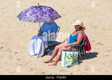 Bournemouth, Dorset, UK. 27th June 2018. UK weather: sunseekers head to the beaches at Bournemouth on another lovely warm sunny day with unbroken blue skies and sunshine. A nice cooling breeze today makes the heat more bearable. Two mature women sitting on the beach, one enjoying the sunshine, the other sitting under parasol with lavender and bees design. Stock Photo
