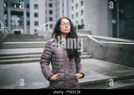Beautiful young woman of European ethnicity with long brunette hair, wearing glasses and a coat stands against backdrop of a business center in the fa Stock Photo