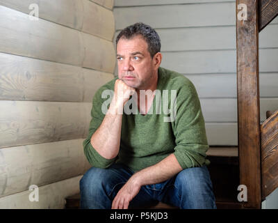 Sad Caucasian middle-aged man sitting on the stairs inside the house Stock Photo