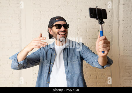 young attractive man in casual clothes hat hipster style holding selfie stick mobile phone shooting self portrait photo or recording video in internet Stock Photo