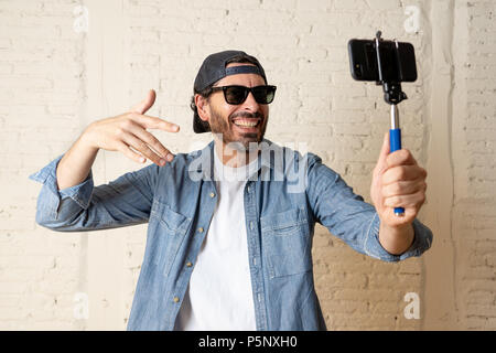 young attractive man in casual clothes hat hipster style holding selfie stick mobile phone shooting self portrait photo or recording video in internet Stock Photo