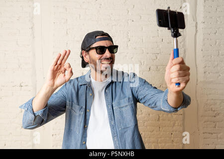 young attractive man in casual clothes hat hipster style holding selfie stick mobile phone shooting self portrait photo or recording video in internet Stock Photo