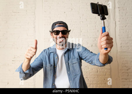 young attractive man in casual clothes hat hipster style holding selfie stick mobile phone shooting self portrait photo or recording video in internet Stock Photo