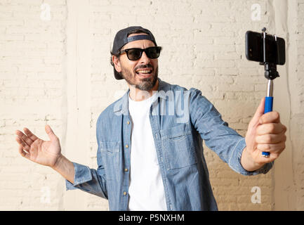 young attractive man in casual clothes hat hipster style holding selfie stick mobile phone shooting self portrait photo or recording video in internet Stock Photo