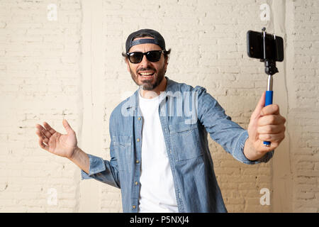 young attractive man in casual clothes hat hipster style holding selfie stick mobile phone shooting self portrait photo or recording video in internet Stock Photo