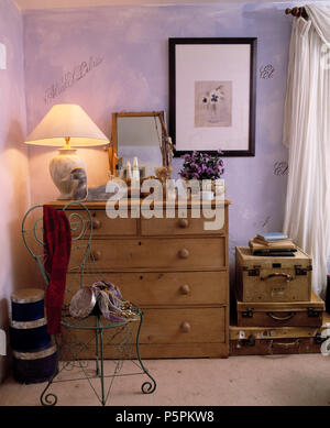 Metal chair and antique pine chest of drawers with lighted lamp in mauve cottage bedroom with stack of old leather suitcases Stock Photo