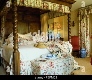 Floral-upholstered ottoman below oak four poster bed with white linen and rose-patterned cushions and bedcover Stock Photo