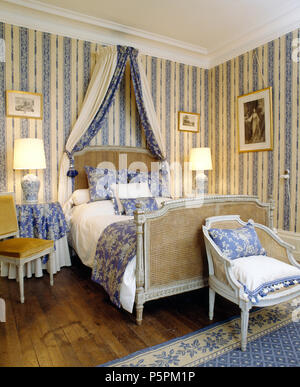 Antique Bergere bed with blue drapes in French country bedroom with blue+white striped wallpaper and lighted lamps Stock Photo