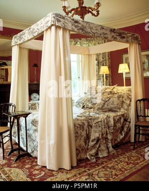 Toile-de-Jouy patterned bed-cover and pillows on four-poster bed with cream drapes in country bedroom with lighted lamps behind bed Stock Photo