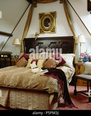 Country bedroom with ornate gilt-framed picture above carved wood antique bed with patterned beige bedlinen and red velvet throw