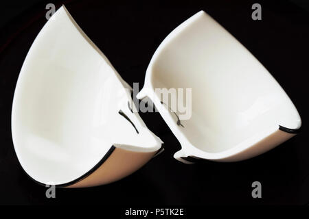 View of a white broken cup and fractured shards of sharp glass fragments scattered on black background. Concept for divorce, relationships, friendship Stock Photo