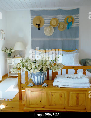 White coastal bedroom with collection of straw hats on pale blue linen wall-hanging above bed with white pillows and linen Stock Photo