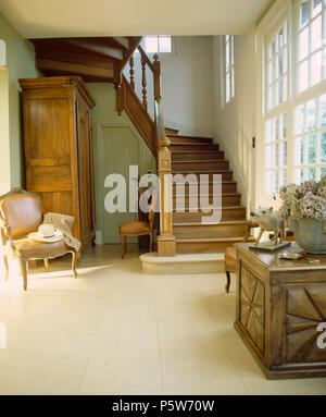 Antique chair and carved woooden chest in French country hall with stone tiled floor and oak staircase Stock Photo