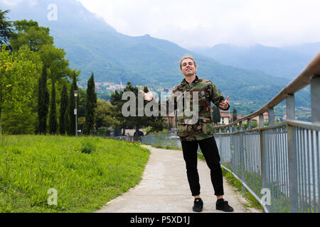 Young caucasian male tourist wearing camouflage jacket standing near banister, lake Como and Alps mountain in background. Stock Photo
