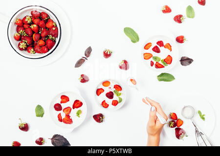 Female's hand holding spoon over a bowl of strawberry ice cream among red berries, mint and basil leaves, plates with desserts, top view. Flat lay com Stock Photo