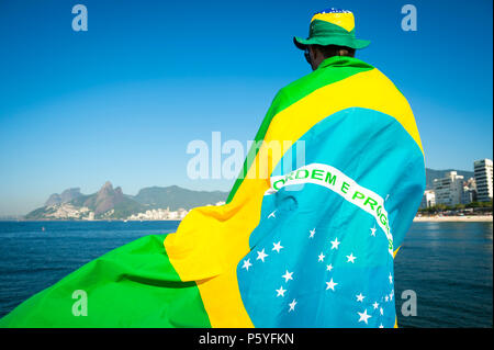 Brazilian football fan wrapped in giant flag and hat in front of Ipanema Beach city skyline Rio de Janeiro. Stock Photo