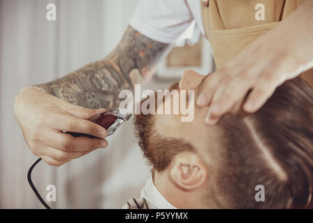 The barber's hand holds hair clipper while cutting the beard. Photo in vintage style