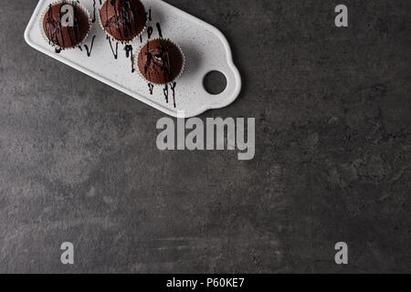 Flat lay of three chocolate muffins topped with chocolate dressing on white serving plate on black concrete background with copy space. Top view. Stock Photo