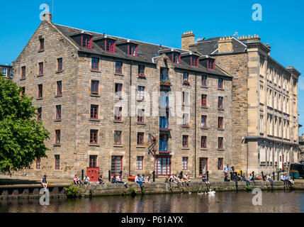 Water of Leith, Edinburgh, Scotland, United Kingdom, 28th June 2018. Office workers lined up sitting on the riverbank in front of converted warehouse into flats called The Cooperage apartments at lunchtime in the hot sunshine during the heatwave with swans and cygnets hoping to be fed Stock Photo