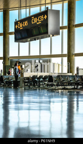 A view of empty seats at boarding area of airport terminal, alongside large glass walls with reflective floors and a solitary man looking out window Stock Photo