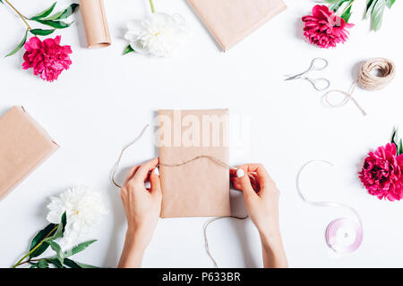 Floral flat lay arrangement woman packing gifts among blooming pink flowers peonies. Top view female hands tying rope on a present. Stock Photo