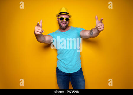 Happy male in blue shirt, sunglasses and yellow hat celebrating success standing with open mouth smiling and pointing fingers of stretched out arms forward on yellow background Stock Photo