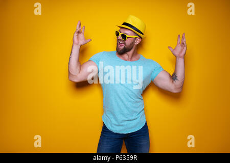 Happy male in blue shirt, sunglasses and yellow hat celebrating success posing with fists up standing with turned aside head over bright yellow studio background Stock Photo