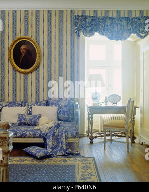 Blue+white floral patterned cushions on sofa with matching throw beside window with festoon blind in living room with striped wallpaper Stock Photo