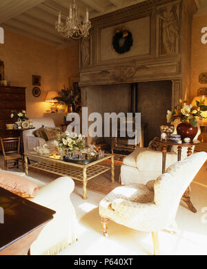 Cream armchair and sofas in French country living room with wood-burning stove in large ornate stone fireplace Stock Photo