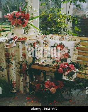 Rose patterned tablecloth and cushion on small table and wooden bench in conservatory Stock Photo