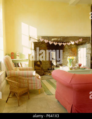 Red sofa and pink striped armchair with small wicker chair in country living room with heart garland on inglenook fireplace Stock Photo