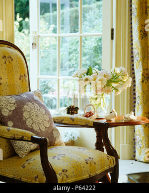 Victorian armchair upholstered in pale yellow Pierre Frey fabric beside small antique table in front of French windows Stock Photo