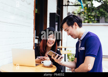One man and woman showing a mobile phone for each other to see with coffee and laptop at outdoor cafe restaurant Stock Photo