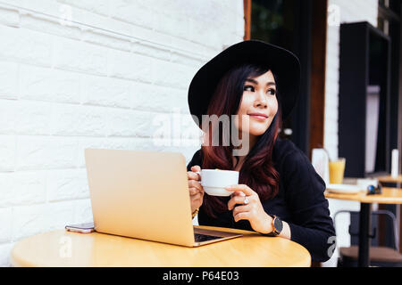 Young fashionable Asian business woman drinking coffee while using a mobile phone and laptop in outdoor cafe restaurant Stock Photo