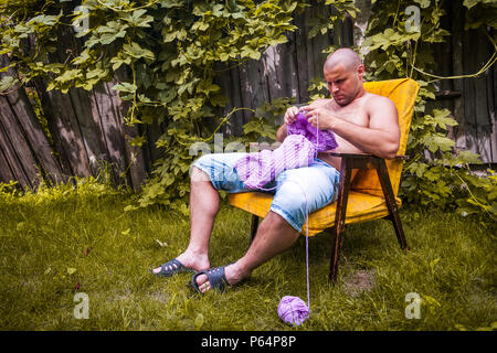 A young bald man sits on an armchair and knits a sweater from natural lilac wool threads at the dacha, in the background a wooden fence, grape bushes Stock Photo