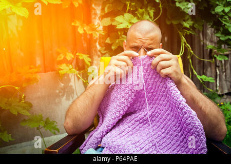 A young bald man sits on an armchair and knits a sweater from natural lilac wool threads at the dacha, in the background a wooden fence, grape bushes Stock Photo