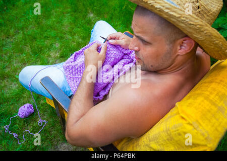 Close-up A young bald man in a straw hat sits on an armchair and knits a sweater from natural lilac woolen threads at the cottage in front of a green  Stock Photo