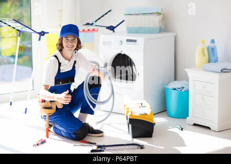 Washing machine repair service. Young male technician in blue uniform examining and repairing broken washer or tumble dryer. Maintenance of home and h Stock Photo