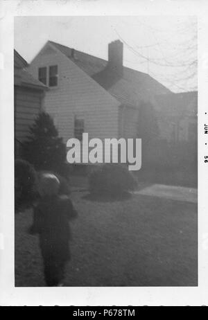 Black and white photograph, slightly out of focus, showing a small, blonde-haired child, moving away from the camera, toward a siding-covered house visible in the background, likely photographed in Ohio, June, 1956. () Stock Photo