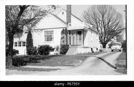 Black and white photograph, showing a blonde woman, wearing sunglasses, and crouching down (presumably to garden) in front of a small, light-colored, tract house, with siding, a covered front porch, and a vintage car at the end of the driveway, likely photographed in Ohio in the decade following World War II, 1950. () Stock Photo