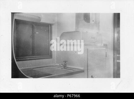 Black and white photograph, showing a small, partially tiled kitchenette located in the cramped interior of a Mid-century caravan or trailer home, with a sink, refrigerator, and window, likely photographed in Ohio in the decade following World War II, 1950. ()