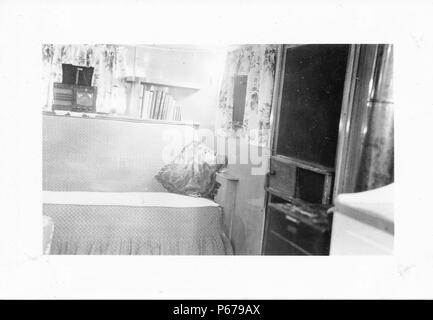 Black and white photograph, showing the cramped interior of a Mid-century caravan or trailer home, with a sofa, bookshelves, a vintage radio, and a door visible at right, likely photographed in Ohio in the decade following World War II, 1950. ()