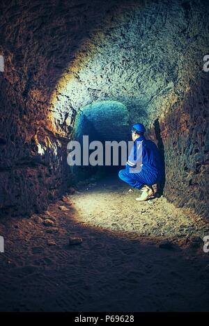 Dry sandstone tunnel, man worker in protective suite in underground. Mysterious dungeon tunnel with walls made inorange sandstone rock Stock Photo